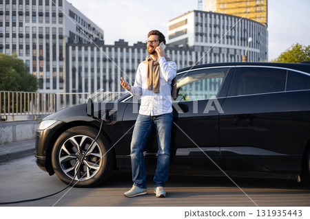 A man talks on his phone while his electric vehicle charges at a charging station in an urban setting. 131935443