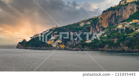 Aerial view of Amalfi, Italy, featuring colorful hillside buildings, steep cliffs, and the Tyrrhenian Sea under a dramatic sunset sky. 131935445