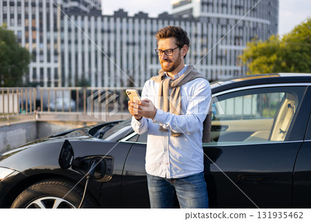 A man checks his phone while his electric car charges at a charging station in an urban setting. A man checks his phone while his electric car charges at a charging station in an urban setting. 131935462