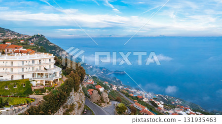 Aerial view of Amalfi, Italy, featuring a white hotel on a cliff, terraces with greenery, coastal road, red roofed buildings, and the Tyrrhenian Sea. Aerial view of Amalfi, Italy, featuring a white hotel on a cliff, terraces with greenery, coastal road, red roofed buildings, and the Tyrrhenian Sea. 131935463