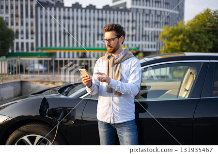 A man checks his phone while his electric vehicle charges at a charging station in an urban setting. A man checks his phone while his electric vehicle charges at a charging station in an urban setting. 131935467
