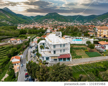 Aerial view of a hillside town in Amalfi, Italy, featuring a white luxury hotel with balconies, a pool, gardens, terracotta roofed houses, and green hills. 131935468