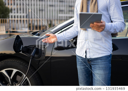A man stands next to his electric vehicle while it charges, holding a tablet. 131935469