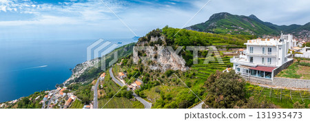Aerial view of the Amalfi Coast in Italy, showing a white hotel on a terraced hillside, lush greenery, cliffs, winding roads, and the Tyrrhenian Sea. 131935473