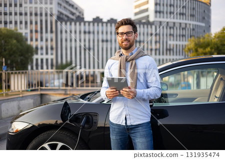 A man stands next to his electric car while it charges, holding a tablet and smiling at the camera. 131935474
