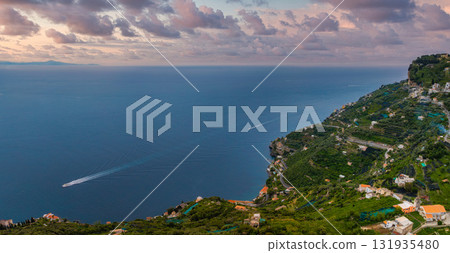 A hillside town with terraced greenery and white buildings overlooks the Tyrrhenian Sea. A boat leaves a trail on calm waters under a colorful sky. 131935480