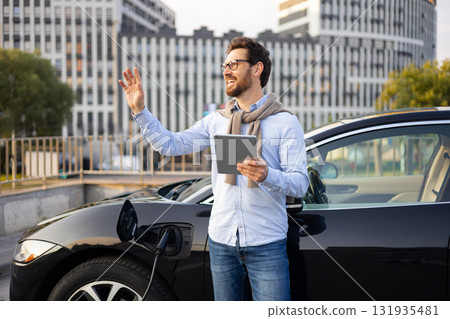 A man waves while standing next to his electric vehicle, which is charging at a station in an urban setting. 131935481