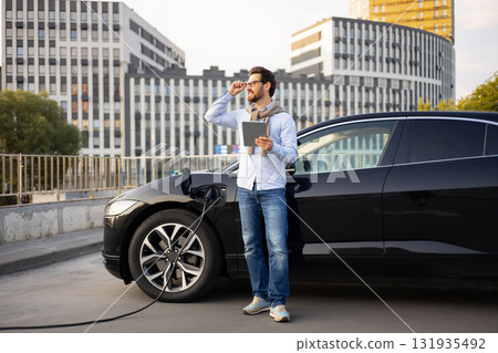 A man stands next to his black electric vehicle while it charges, holding a tablet and looking towards the city. A man stands next to his black electric vehicle while it charges, holding a tablet and looking towards the city. 131935492