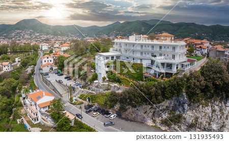 Aerial view of a white ivy covered hotel on a rocky cliff, surrounded by greenery, a winding road, terracotta roofed houses, and rolling hills at sunset. 131935493