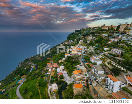 Aerial view of Ravello, Italy, with terracotta roofed buildings, terraced gardens, and the Amalfi Coast under a vibrant sunset sky. 131935502
