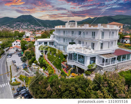 A white multi story hotel on a lush hillside with balconies, surrounded by greenery, a small town, a winding road, and a vibrant sunset sky. A white multi story hotel on a lush hillside with balconies, surrounded by greenery, a small town, a winding road, and a vibrant sunset sky. 131935516