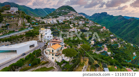 Aerial view of Ravello, Italy, featuring white and pastel buildings, terraced gardens, a modern structure, and lush green mountains under a cloudy sky. Aerial view of Ravello, Italy, featuring white and pastel buildings, terraced gardens, a modern structure, and lush green mountains under a cloudy sky. 131935517