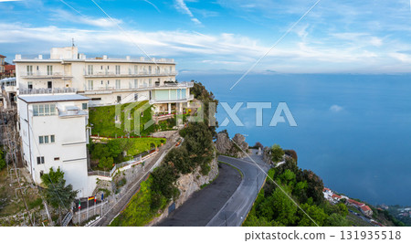 Aerial view of a white luxury hotel with balconies and green terrace on a cliff in Amalfi, Italy, overlooking the Tyrrhenian Sea and winding road. Aerial view of a white luxury hotel with balconies and green terrace on a cliff in Amalfi, Italy, overlooking the Tyrrhenian Sea and winding road. 131935518