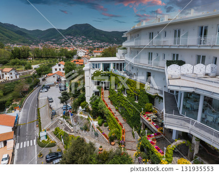 Luxurious hotel with ivy covered facade and terraces on a hillside in Amalfi, Italy, surrounded by greenery, houses, and rolling hills under a colorful sky. 131935553