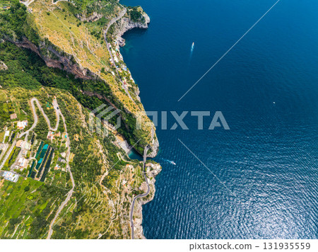 Aerial image of the Amalfi Coast, Italy, featuring cliffs, winding roads, terraced fields, green hills, and a boat on the Tyrrhenian Sea. 131935559