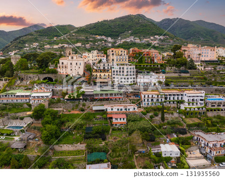 Aerial view of Ravello, Italy, featuring hillside terraces, historic buildings in warm tones, lush greenery, and a vibrant orange and pink sunset sky. Aerial view of Ravello, Italy, featuring hillside terraces, historic buildings in warm tones, lush greenery, and a vibrant orange and pink sunset sky. 131935560