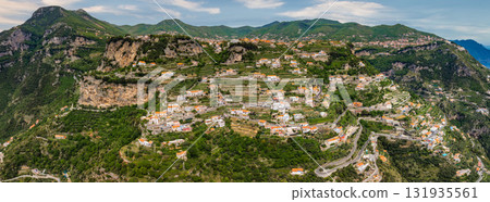 Aerial view of Amalfi, Italy, showcasing terraced landscapes, pastel colored buildings, lush greenery, steep cliffs, and winding roads on a hillside. 131935561