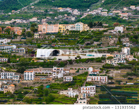 Aerial view of Ravello, Italy, featuring the white Auditorium Oscar Niemeyer, terraced gardens, colorful villas, and lush greenery on a hillside. 131935562