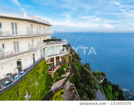 A modern white luxury hotel with balconies sits on a cliff in Amalfi, Italy, surrounded by greenery, with a winding road and the Tyrrhenian Sea below. A modern white luxury hotel with balconies sits on a cliff in Amalfi, Italy, surrounded by greenery, with a winding road and the Tyrrhenian Sea below. 131935570