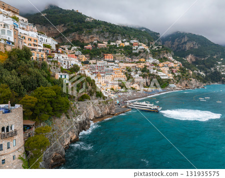 The coastal town of Positano, Italy, features cascading colorful buildings, a ferry near the shore, lush greenery, and low clouds over rugged cliffs. The coastal town of Positano, Italy, features cascading colorful buildings, a ferry near the shore, lush greenery, and low clouds over rugged cliffs. 131935575