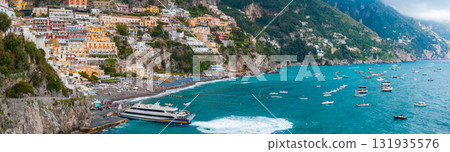 Positano on Italy's Amalfi Coast features vibrant buildings on steep cliffs, a busy beach with umbrellas, a ferry, and boats in turquoise waters. 131935576
