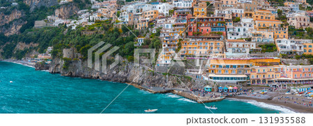 View of Positano on the Amalfi Coast, with vibrant buildings on a steep hillside, a beach, boats, turquoise waters, and surrounding greenery. 131935588
