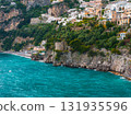 The coastal town of Positano, Italy, features pastel buildings on steep cliffs, a stone watchtower, turquoise waters, and lush greenery along the Amalfi Coast. 131935596