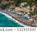 A vibrant beach in Positano, Italy, with colorful umbrellas, turquoise waters, and pastel Mediterranean buildings set against steep, green cliffs. 131935597