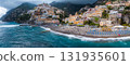 View of Positano's stacked hillside buildings, a church dome, sandy beach with blue umbrellas, turquoise sea, cliffs, and lush greenery under a cloudy sky. 131935601