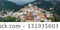View of Positano's vibrant buildings in pastel hues stacked on a steep hillside, surrounded by greenery and cloud covered mountains in the background. 131935603
