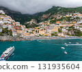 The coastal town of Positano, Italy, features cascading colorful buildings, a beach, boats on the sea, and lush mountains with low hanging clouds. 131935604
