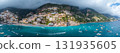 Positano's vibrant buildings cascade down steep cliffs to the Mediterranean Sea, with boats, a beach lined with umbrellas, and lush mountains in view. 131935605