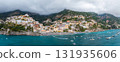 Positano on Italy's Amalfi Coast features vibrant buildings on steep hills, turquoise waters with boats, lush green hills, and low hanging clouds. 131935606