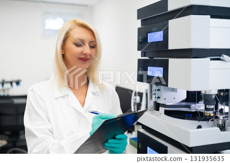 Senior female scientist in a white lab coat making detailed notes during HPLC analysis in a modern chemical laboratory. Concept of professional research, precise data documentation, and advanced 131935635