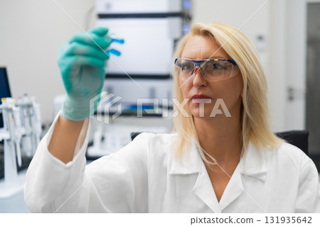 Female scientist in lab coat observing blue biochemical solution in test tube for research purposes. 131935642