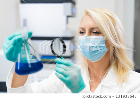 Female chemist wearing protective mask in laboratory, carefully inspecting chemical solution in flask using magnifying glass. Female chemist wearing protective mask in laboratory, carefully inspecting chemical solution in flask using magnifying glass. 131935643