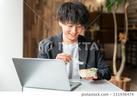 A young Asian male businessman eating a lunch box while looking at a laptop in an eat-in space or company cafeteria A young Asian male businessman eating a lunch box while looking at a laptop in an eat-in space or company cafeteria 131936474
