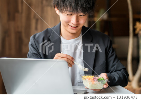 A young Asian male businessman eating a lunch box while looking at a laptop in an eat-in space or company cafeteria 131936475