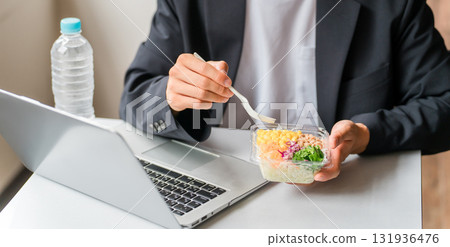 A young Asian male businessman eating a lunch box while looking at a laptop in an eat-in space or company cafeteria 131936476