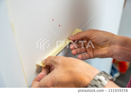 Man covering a light switch with masking tape before painting the wall white. Home renovation, surface protection and careful preparation during interior work. 131939356