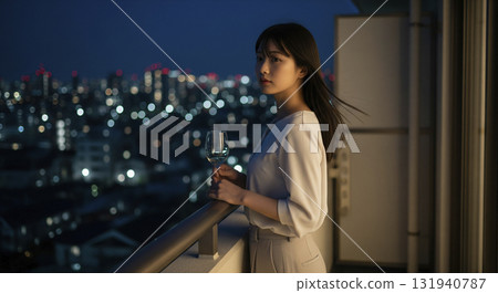 一名女子站在公寓大樓的陽台上，凝視著夜景。她手捧酒杯，長髮在夜風中飄揚。 131940787