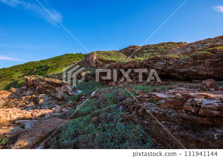Close-up of rugged red and brown coastal rocks at Khao Laem Ya, Rayong, Thailand, contrasting with the bright blue sky and lush green slopes 131941144
