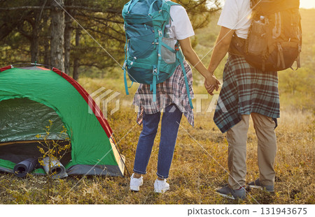 Couple hikers with backpacks holding hands, tourists standing by tent at sunset Couple hikers with backpacks holding hands, tourists standing by tent at sunset 131943675