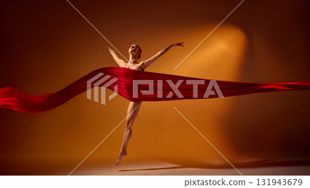 Ballet dancer in red fabric leaping joyfully under orange stage lighting 131943679