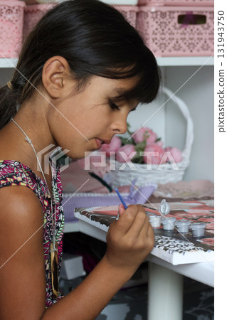 A girl paints a picture using paints and a brush. Paint by numbers. A fragment of the canvas and paint containers are visible. Side Profile Shot 131943750