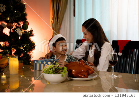Happy family celebrating Christmas together at home with gifts and dinner. Smiling mother and son enjoying festive holiday moments with decorated tree and warm lighting atmosphere full of love and joy Happy family celebrating Christmas together at home with gifts and dinner. Smiling mother and son enjoying festive holiday moments with decorated tree and warm lighting atmosphere full of love and joy 131945165