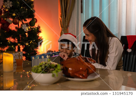 Curious boy opening Christmas gift box with his loving mother beside decorated tree at home. Warm holiday atmosphere with festive dinner, candlelight, and joyful family celebration full of excitement Curious boy opening Christmas gift box with his loving mother beside decorated tree at home. Warm holiday atmosphere with festive dinner, candlelight, and joyful family celebration full of excitement 131945170