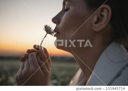 A woman is holding a flower and smelling it. The image has a peaceful and calming mood, as the woman is enjoying the simple pleasure of smelling the flower. A woman is holding a flower and smelling it. The image has a peaceful and calming mood, as the woman is enjoying the simple pleasure of smelling the flower. 131945714