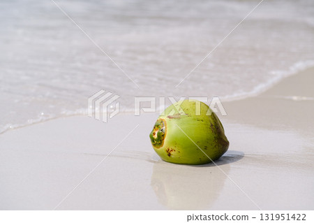 Green coconut rests on a sandy beach as waves gently lap at the shore under a clear sky on a sunny day 131951422