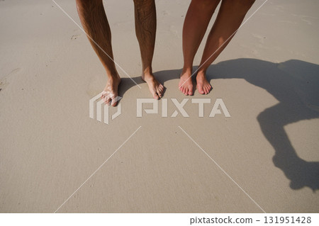 Couple standing barefoot on sandy beach with sunlit shoreline and gentle waves during a warm summer day Couple standing barefoot on sandy beach with sunlit shoreline and gentle waves during a warm summer day 131951428